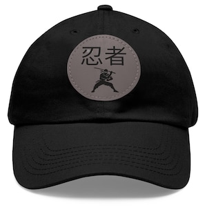 Peut inclure: Casquette de baseball noire avec une visière incurvée. Un écusson circulaire sur le devant présente des caractères japonais et une silhouette de ninja. Un accessoire décontracté.