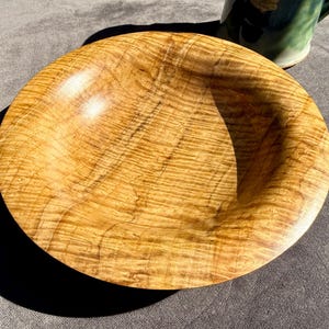 Puede incluir: Un cuenco de madera hecho a mano con un acabado marrón claro natural. El cuenco tiene un diseño ancho y poco profundo y una superficie lisa y pulida. La veta de la madera es visible, lo que añade encanto rústico al cuenco. Una pieza decorativa para el hogar.