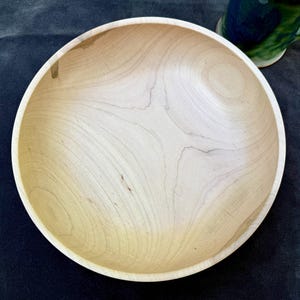 May include: A light-colored, round wooden bowl with a smooth, polished surface. The bowl has a wide, shallow design and a natural wood grain pattern. The bowl is likely handmade and suitable for serving food or as a decorative item.