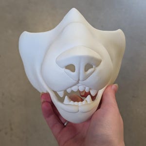 May include: A white, unpainted fursuit mask of a canine face, featuring a prominent nose, open mouth with visible teeth, and a detailed design. The mask is held by a hand.