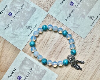 May include: A turquoise and white beaded bracelet with a silver dreamcatcher charm. The bracelet is displayed on a wooden surface with three cards featuring zodiac signs and descriptions. The cards read "Pisces Mystic Intuition", "Cancer Lunar Serenity", and "Scorpio Shadows Transform".