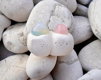 May include: Two white ceramic figures with faces, each holding a crystal. One crystal is light blue and the other is pink. The figures are sitting on a bed of white stones.