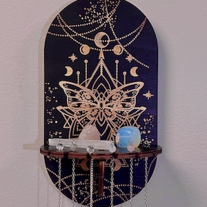 Op de afbeelding: Een houten wanddecoratie met een halve maan en vlinderontwerp. De decoratie heeft een plank om kristallen te tonen en een set kettingen die aan de onderkant hangen.
