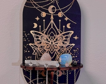 May include: A wooden wall hanging with a crescent moon and butterfly design. The hanging has a shelf for displaying crystals and a set of chains hanging from the bottom.