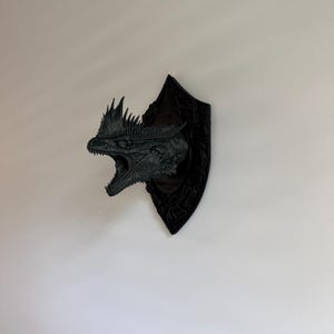 May include: A black wall-mounted dragon head sculpture featuring a detailed, open-mouthed expression with sharp teeth and a spiky crest. The dragon head is attached to a diamond-shaped black plaque with intricate, embossed patterns on its surface.