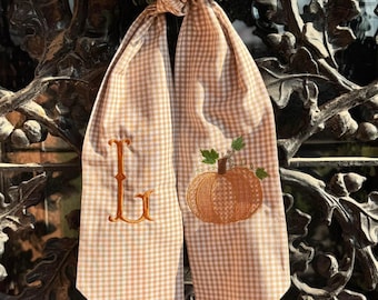 May include: A tan and white gingham scarf with an embroidered orange pumpkin and the letter "L". The scarf is tied in a knot and hangs in front of a green wreath and a wrought iron gate.