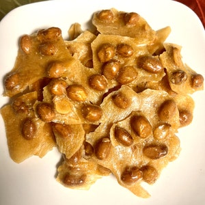 May include: A close-up of a plate of peanut brittle, broken into irregular pieces. The brittle is a light golden brown color and is covered in peanuts.