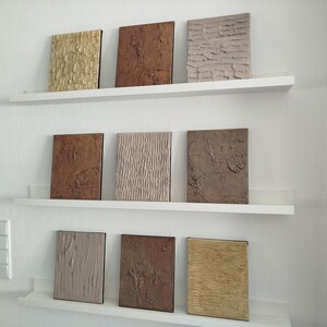 May include: Nine textured square wall art pieces in shades of brown, beige, and gold are displayed on white shelves against a white wall. The art pieces have various surface patterns, including rough, ridged, and smooth textures.