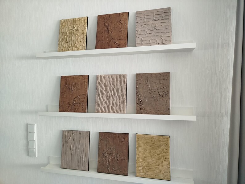 May include: Nine textured square wall art pieces in shades of brown, beige, and gold are displayed on white shelves against a white wall. The art pieces have various surface patterns, including rough, ridged, and smooth textures.