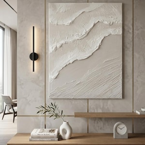 Könnte beinhalten: Abstrakte, strukturierte Wandkunst in neutralen Tönen mit einem geschichteten Design. Das Kunstwerk hängt über einem Holzschrank mit einer weißen Vase, Büchern und einer kleinen Uhr. Eine schwarze Wandleuchte ist an der Wand befestigt.