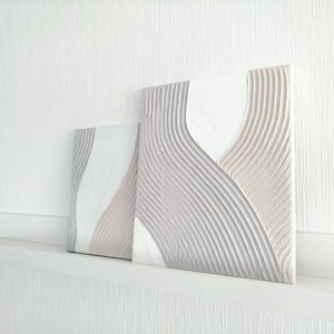 Könnte beinhalten: Zwei abstrakte, strukturierte Wandkunstwerke. Das Kunstwerk weist eine neutrale Farbpalette mit Weiß- und Beigetönen auf. Die Designs haben ein wellenartiges Muster, das ein Gefühl von Bewegung und Tiefe erzeugt. Die Stücke werden auf einem weißen Regal ausgestellt.