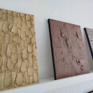May include: Three textured wall art panels in neutral tones. The first panel is light beige with a repeating pattern. The second panel is a reddish-brown, and the third is gray. All panels have a black border.