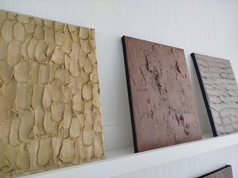 May include: Three textured wall art panels in neutral tones. The first panel is light beige with a repeating pattern. The second panel is a reddish-brown, and the third is gray. All panels have a black border.