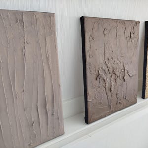 May include: Three textured canvas art pieces in neutral tones. The first is a light beige with vertical strokes, the second is a darker taupe with a more abstract texture, and the third is a light gold with a wood grain pattern.