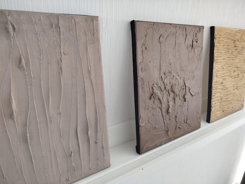 May include: Three textured canvas art pieces in neutral tones. The first is a light beige with vertical strokes, the second is a darker taupe with a more abstract texture, and the third is a light gold with a wood grain pattern.