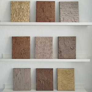 May include: Nine textured abstract art panels in various earth tones, including beige, brown, and tan, are displayed on white shelves. Each rectangular panel features a unique surface design, creating a visually interesting display of textures and colors.