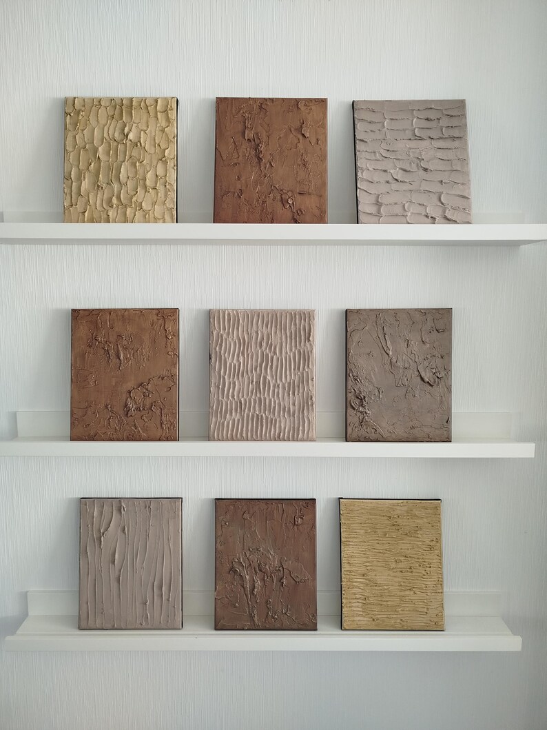 May include: Nine textured abstract art panels in various earth tones, including beige, brown, and tan, are displayed on white shelves. Each rectangular panel features a unique surface design, creating a visually interesting display of textures and colors.