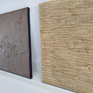 May include: Three textured abstract art pieces. The artwork features neutral tones of beige, gray, and white with black edges. The textures appear to be created with a thick application of paint or plaster, creating a tactile surface. The pieces are displayed on a white shelf.