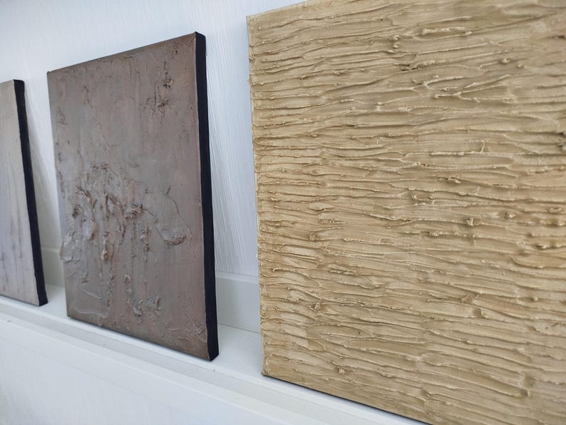 May include: Three textured abstract art pieces. The artwork features neutral tones of beige, gray, and white with black edges. The textures appear to be created with a thick application of paint or plaster, creating a tactile surface. The pieces are displayed on a white shelf.