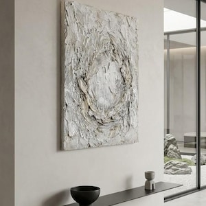 Könnte beinhalten: Ein quadratisches Wandbild mit dicker, geschichteter weißer und beiger Impasto-Textur und einem zentralen kreisförmigen Motiv. Unter dem Bild befindet sich ein schmales, schwarzes Wandregal mit einer mattschwarzen Schale und einem kleinen grauen Skulpturobjekt.