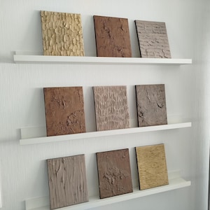 May include: Nine textured wall art panels in various shades of brown, beige, and tan are displayed on white shelves. The panels have unique surface patterns, including vertical lines, rough textures, and organic shapes. The arrangement is on a white wall.