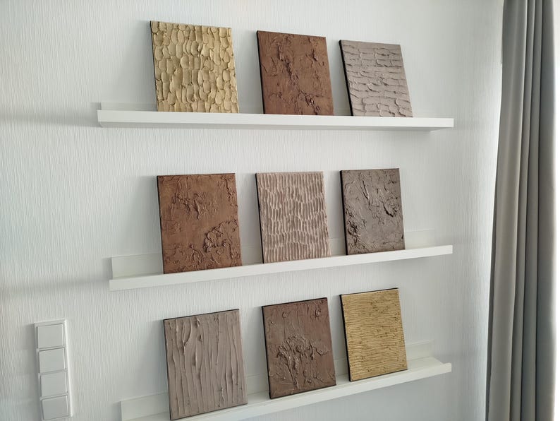 May include: Nine textured wall art panels in various shades of brown, beige, and tan are displayed on white shelves. The panels have unique surface patterns, including vertical lines, rough textures, and organic shapes. The arrangement is on a white wall.