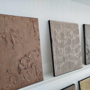 May include: A collection of textured abstract art panels in various earth tones. The panels are rectangular, with raised, sculpted surfaces. The colors range from a deep brown to a light beige, with black edges. The art is displayed on a white shelf.