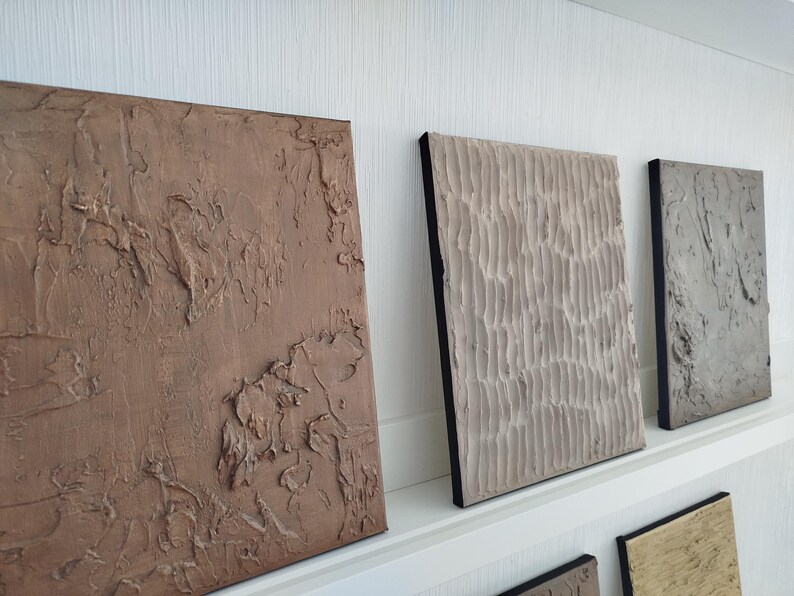 May include: A collection of textured abstract art panels in various earth tones. The panels are rectangular, with raised, sculpted surfaces. The colors range from a deep brown to a light beige, with black edges. The art is displayed on a white shelf.