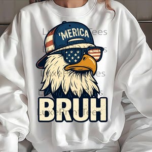 以下が含まれることがあります： 白いスウェットシャツ。サングラスと「'MERICA」の文字が入った帽子をかぶったワシのグラフィック。ワシのサングラスは旗のデザインで、下に「BRUH」の文字が入っています。