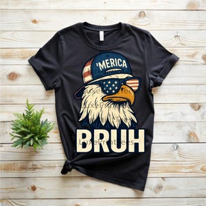 以下が含まれることがあります： サングラスと「'MERICA」の文字が入った帽子をかぶったワシのグラフィックが特徴の黒いTシャツ。「BRUH」の文字が太字で下にプリントされています。