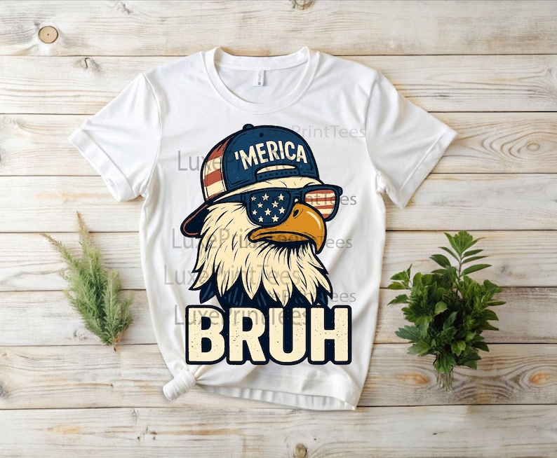 以下が含まれることがあります： サングラスと「'MERICA」の文字が入った帽子をかぶった漫画のワシが描かれた白いTシャツ。ワシは愛国的なデザインです。「BRUH」の文字が下に印刷されています。Tシャツは木製の背景にあります。