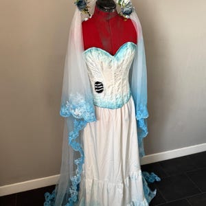 May include: A white and blue bridal gown with a corset-style bodice and a long skirt. The dress features a blue lace veil and a floral headpiece. The gown is displayed on a mannequin, showcasing its design and details. The dress has a distressed look.
