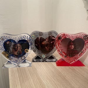 Peut inclure: Trois cadres photo en forme de cœur avec des accents de paillettes. Les cadres sont transparents avec une finition pailletée argentée, bleue et rouge. Chaque cadre présente une image différente d'une personne.