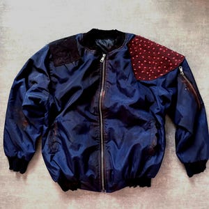Puede incluir: Una chaqueta bomber azul marino con cierre de cremallera. La chaqueta tiene un parche negro en el hombro izquierdo y un parche rojo con pequeños puntos blancos en el hombro derecho.
