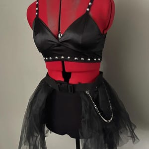 May include: A black satin crop top with silver studs and a black choker with a silver heart pendant. The top is paired with a black tulle skirt with a silver chain and a black belt.