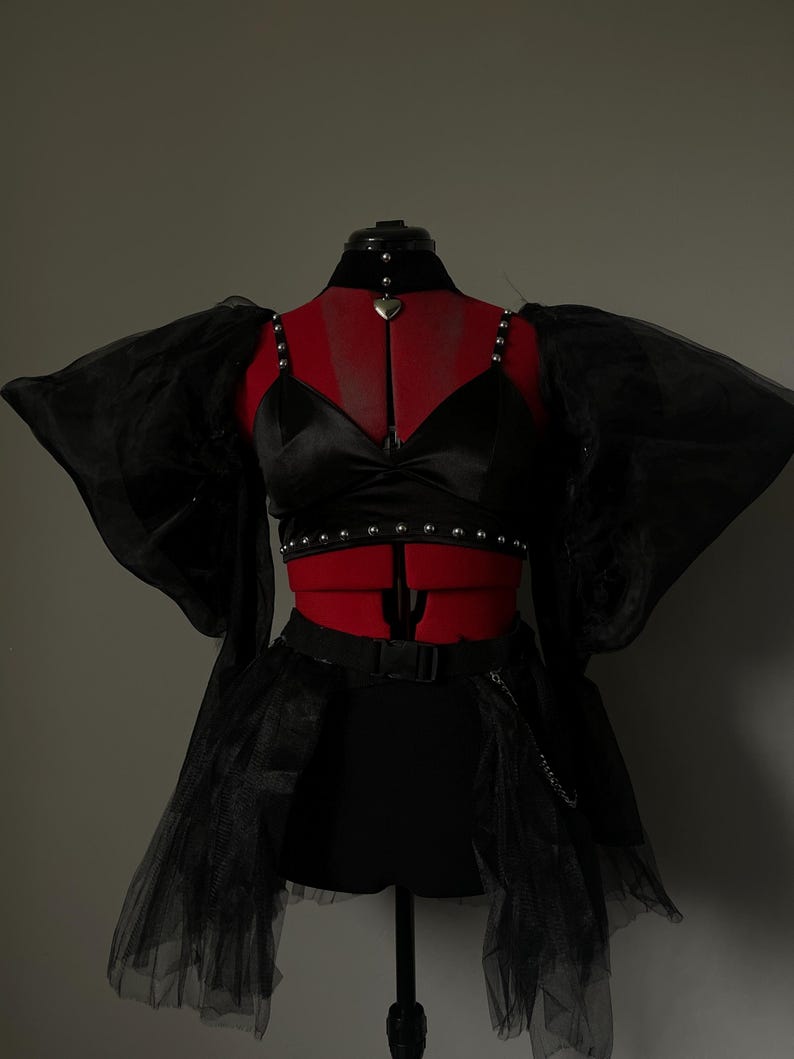 May include: A black satin crop top with a heart-shaped pendant necklace and a black tulle skirt with a chain belt. The top has a beaded trim and the skirt has a layered design.