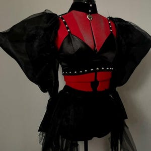 May include: A black crop top with puff sleeves and a black tulle skirt. The top has a satin bodice with silver studs and a silver heart pendant necklace.