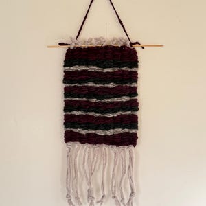 May include: A burgundy and gray woven wall hanging with a fringe of white yarn. The hanging is attached to a wooden dowel rod with a string.