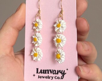 May include: A pair of dangle earrings on a light pink display card labeled Lunvary Jewelry Co. Each earring features three white daisy flowers with yellow centers connected by gold-toned metal jump rings, hanging from gold-toned earring hooks.