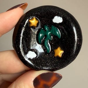 Könnte beinhalten: Ein runder, schwarzer Harzanhänger mit Glitzer. Der Anhänger zeigt einen grünen Drachen, zwei gelbe Sterne und zwei weiße Wolken. Der Anhänger wird in einer Hand mit einem Schildpatt-Nageldesign gehalten.