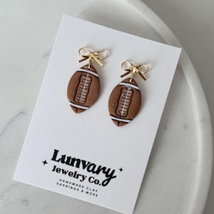 Puede incluir: Pendientes marrones con forma de balón de fútbol con costuras blancas y lazos dorados. Se muestran en una tarjeta blanca con el texto "Lunvary Jewelry Co." y "HANDMADE CLAY EARRINGS & MORE".