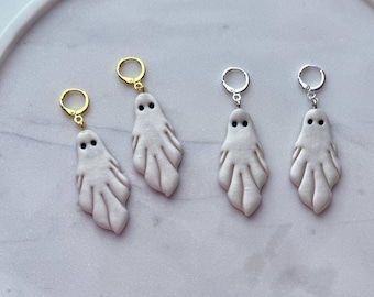 May include: Four ghost-shaped earrings with black dot eyes and white bodies. Two earrings have gold-coloured hoops, and two have silver-coloured hoops. The earrings are displayed on a white and grey marble-patterned tray.