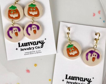 Könnte beinhalten: Zwei Paar Hängeohrringe auf weißen Präsentationskarten mit der Aufschrift Lunvary Jewelry Co. Die Ohrringe bestehen aus runden, beigen Tonanhängern mit orangefarbenen Kürbissen und violetten Geistern mit orangefarbenen Mündern.