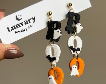 May include: Halloween-themed earrings spelling out "BOO". The earrings feature black and white letters with ghost and bat charms, and orange letter "O"s. The earrings are made of polymer clay and have gold-colored ear wires. The brand name "Lunvary Jewelry Co." is visible.