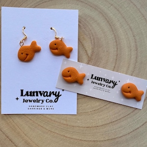 May include: Orange goldfish-shaped earrings with gold-coloured hooks. The earrings are displayed on a white card with the text "Lunvary Jewelry Co." and "Handmade Clay Earrings & More." A matching charm is also shown.