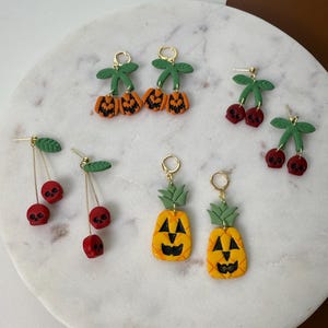 May include: A collection of handmade Halloween-themed earrings. The earrings feature designs such as jack-o'-lanterns, cherries with skull faces, and pineapples with jack-o'-lantern faces. The earrings are made of clay and have gold-colored earring hooks.