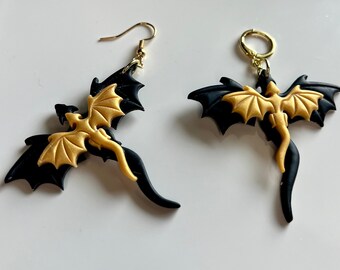 May include: A pair of black and gold dragon earrings. The dragons are in flight with their wings spread. The earrings are made of polymer clay.