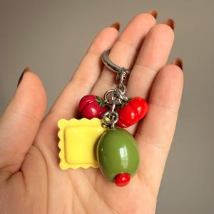 Puede incluir: Un llavero plateado con colgantes temáticos de comida. Los colgantes incluyen dos tomates rojos, un ravioli amarillo y una aceituna verde con un punto rojo. Los colgantes están unidos a un anillo plateado.