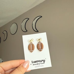May include: A pair of handmade clay earrings shaped like footballs. The brown earrings have white stripes and are topped with gold bows. The earrings are displayed on a white card with the text "Lunvary Jewelry Co."