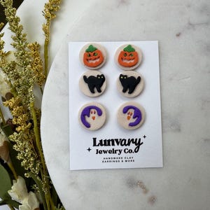 Może przedstawiać: Zestaw sześciu kolczyków sztyftów o tematyce Halloween. Kolczyki przedstawiają wzory dyni, czarnego kota i ducha. Kolczyki znajdują się na białej karcie z napisem "Lunvary Jewelry Co. HANDMADE CLAY EARRINGS & MORE".
